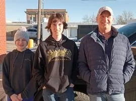 Three people standing outdoors in casual clothing on a sunny day with buildings in the background
