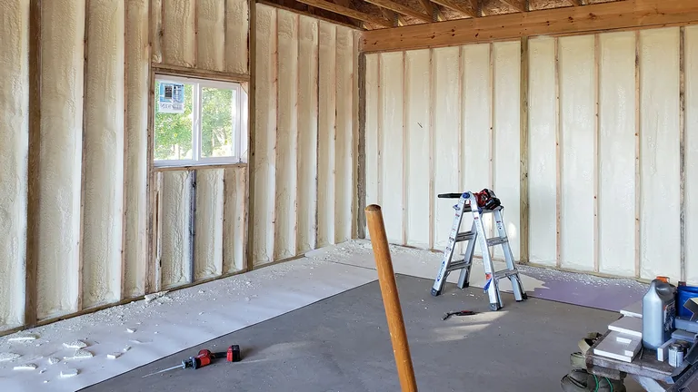Interior room under construction with wood framing, spray foam insulation, window, ladder, and tools scattered on floor