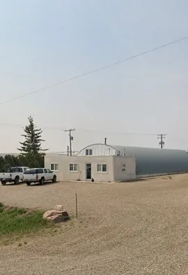 Small white building and large gray industrial structure beside a gravel parking area with two pickup trucks parked nearby