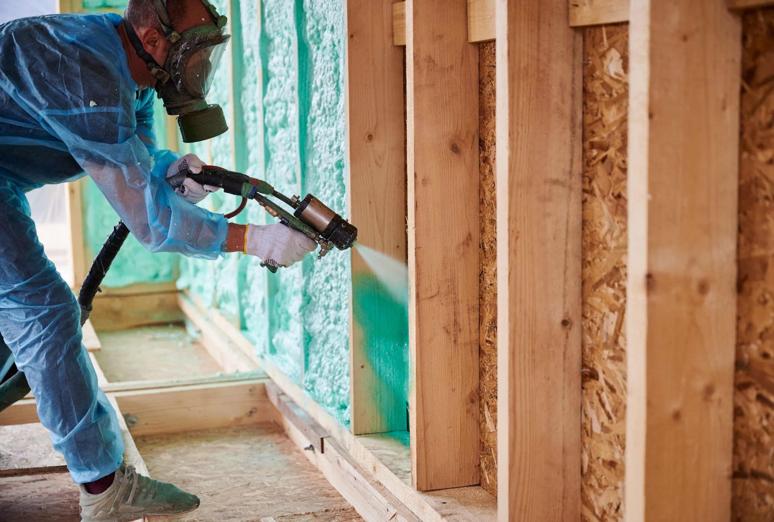 Worker in protective gear spraying polyurethane foam insulation within wooden house frame wall cavity using spray gun