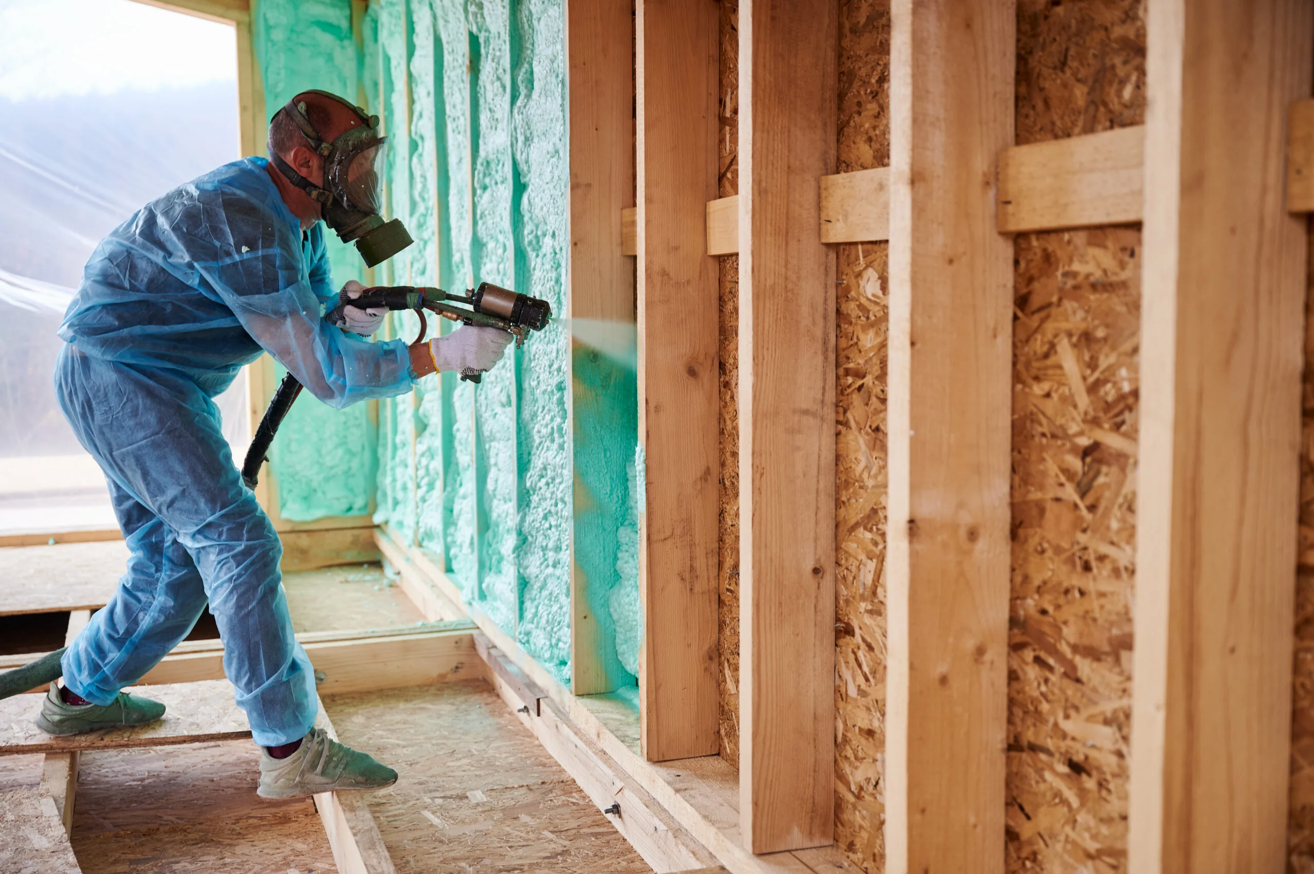 Worker in protective gear spraying green polyurethane foam insulation onto wooden frame interior wall of house under