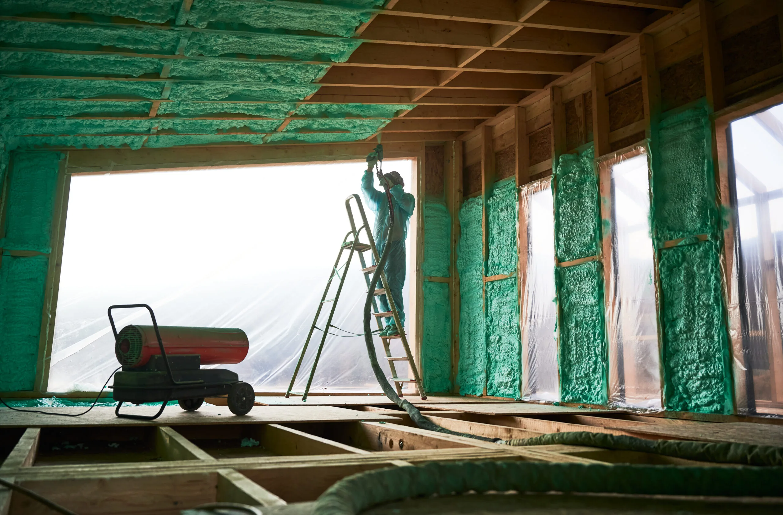 Construction worker on ladder spraying green polyurethane foam insulation on wooden frame interior walls and ceiling.